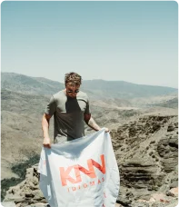 Estudante de camiseta e óculos escuro no topo de uma montanha com bandeira KNN Idiomas