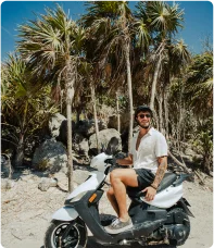 Homem em uma moto branca na areia entre vegetação verde com palmeiras