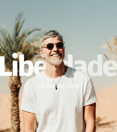 Ator Reynaldo Gianecchini sorrindo de óculos escuro e camiseta branca, em um ambiente aberto com areia e palmeiras no Marrocos, na África
